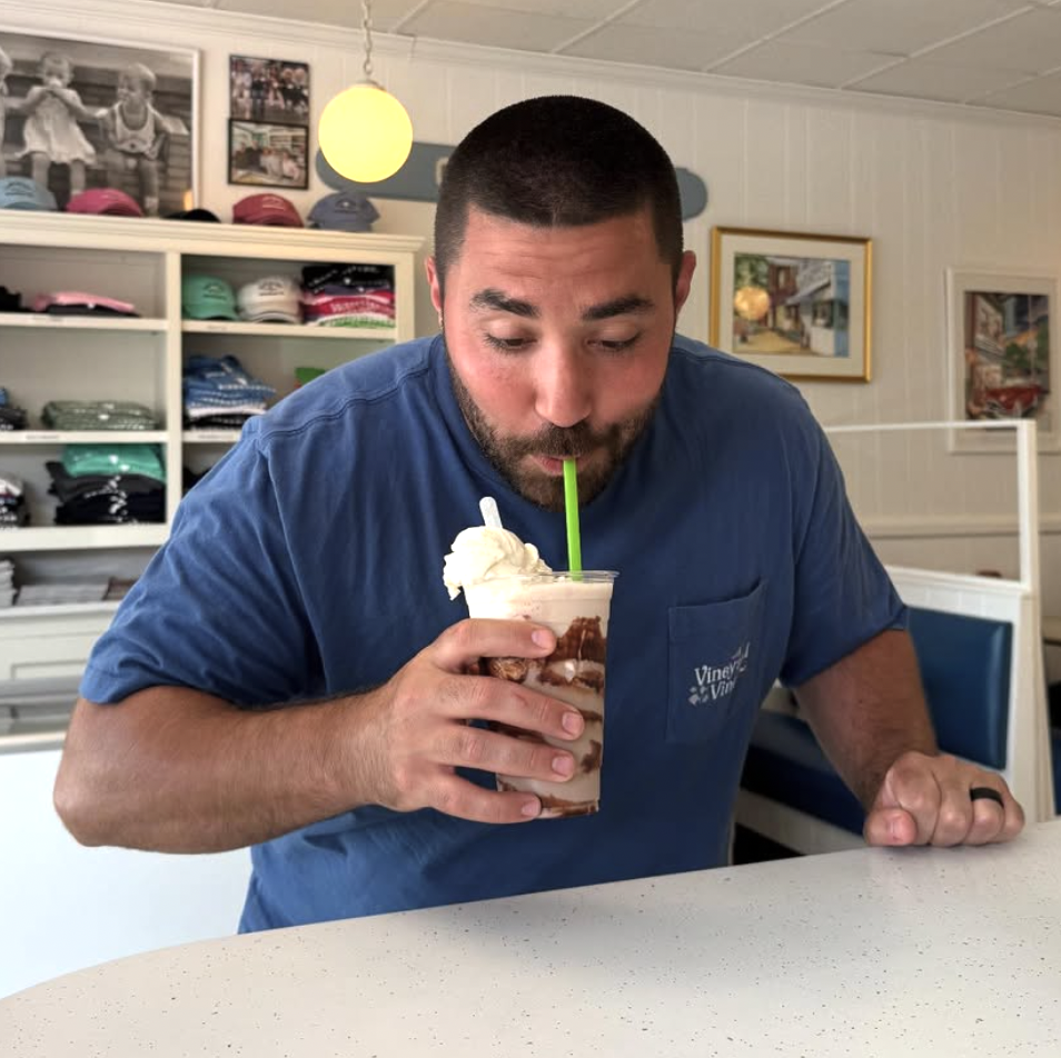 Customer leaning over the counter sipping a towering chocolate milkshake topped with whipped cream